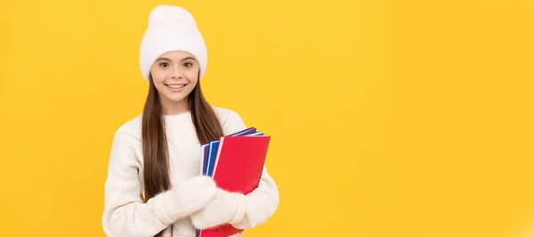 Winter school. happy kid in winter hat and gloves hold homework on yellow background, copy space, back to school. Banner of school girl student. Schoolgirl pupil portrait with copy space