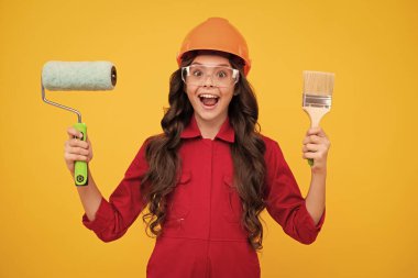 Child builder in hard hat helmet. Teenage girl painter with painting brush tool or paint roller. Teenager worker isolated on yellow background. Kids renovation concept. Excited teenager girl