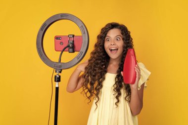 Teen girl speaking in front of camera for vlog isolated on yellow background. Teenager working as blogger hold hair conditioner or shampoo bottle, recording video blog
