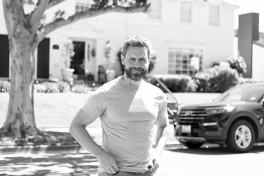 Gülümseyen, mavi gömlekli, sakallı bir adam evin ve arabanın önünde. Keyfi yerinde..