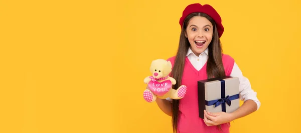 amazed teen girl in french beret hold toy present and gift box on yellow background, amazement. Kid girl with gift, horizontal poster. Banner header with copy space