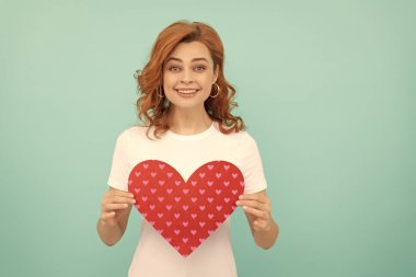 Gülümseyen kızıl saçlı kadın mavi arka planda kırmızı kalbi tutuyor..