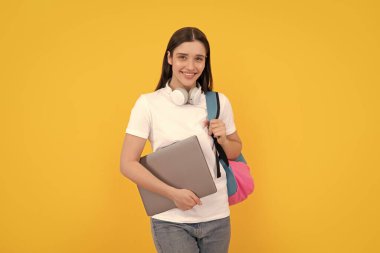 Sırt çantalı genç kız dizüstü bilgisayarını sarı arka planda kulaklıkla tutuyor, eğitim görüyor..