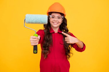 Kasklı çocuk müteahhiti. Fırça aletli ya da boya silindirli genç kız ressam. Genç işçi sarı arka planda izole edilmiş. Çocuk yenileme konsepti. Mutlu gülümseyen genç kız