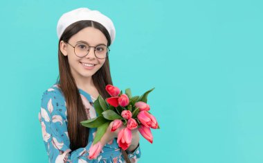 glad child in beret with tulips. mothers or womens day. kid in eyeglasses hold flowers for 8 of march. teen girl with spring bouquet on blue background. floral present. copy space.
