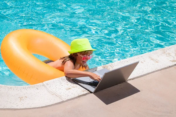 Havuzda rahatlayan bir çocuk, yaz suyunda dizüstü bilgisayar kullanıyor. Çocuk online çalışma ya da tropikal deniz plajında çalışıyor. Yaşam konsepti teknolojisi
