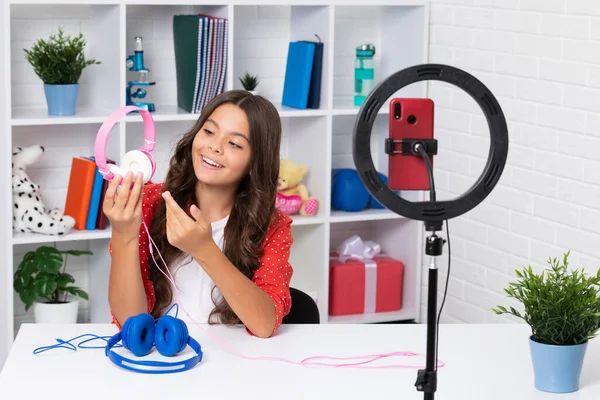 Teenager blogger making video, child vlogger influencer record content. Child blogger records video on mobile phone using ring lamp, video lights for blog or vlog at home. Happy smiling girl