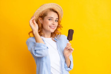 Genç kadın, sarı arka planda çikolata jöleli dondurma yiyor. Yazın hasır şapkalı komik kızıl kız dondurma yalıyor.