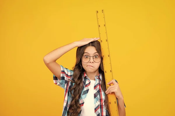 Kid height measure, growth measurement. Funny face. School girl holding measure for geometry lesson, isolated on yellow background. Measuring equipment. Student study math