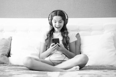 Surprised face, surprise emotions of teenager girl. Teenager child girl wearing headphones watching videos listening music on smart phone sitting on bed in her room
