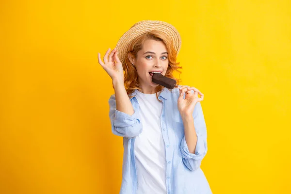 Young woman eat ice creams with chocolate glaze on yellow background. Funny redhead girl with summer straw hat lick ice cream