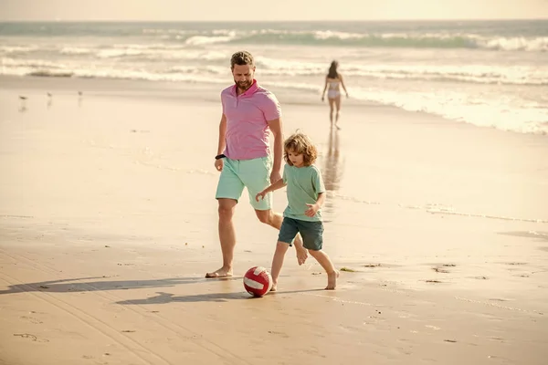 father and son play football on beach. daddy with kid boy on hot summer day. weekend family day. dad and child having fun outdoors. childhood and parenting. family holidays. sport activity.