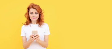 smiling redhead woman chatting in smartphone or make online shopping on yellow background, blogging. Woman isolated face portrait, banner with mock up copy space