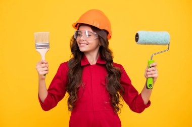 Miğferli çocuk müteahhidi. Fırça aletli ya da boya silindirli genç kız ressam. Sarı arka planda izole edilmiş tamir işindeki çocuk. Çocuk yenileme konsepti. Mutlu genç kız
