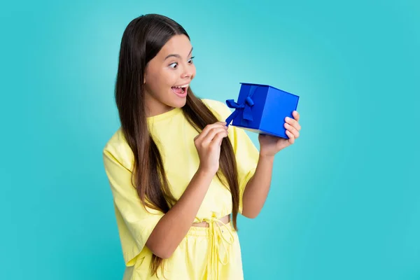 Surprised face, surprise emotions of teenager girl. Teenage child holding gift box on blue isolated background. Gift for kids birthday. Christmas or New Year present box