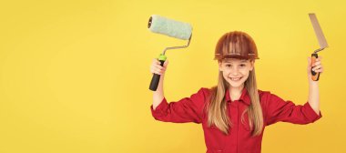 glad teen child in builder hard hat with paint roller and spatula on yellow wall. Child builder in helmet horizontal poster design. Banner header, copy space