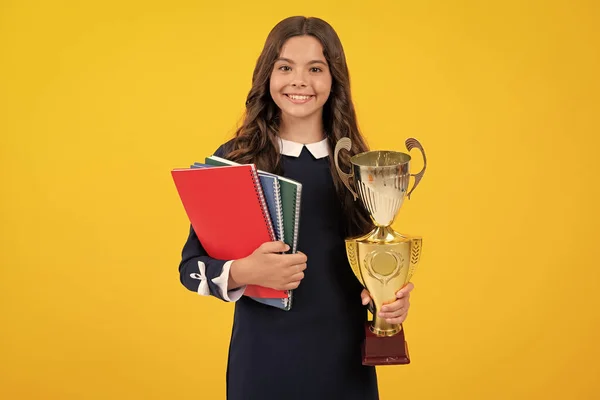 Teenager school girl with award winner trophy. Child hold books with gold trophy or winning cup isolated on yellow. Education graduation, victory and winning