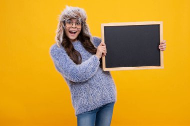 Teenage girl child in warm winter hat and sweater holding blackboard, isolated on a yellow background. Excited face, cheerful emotions of teenager girl