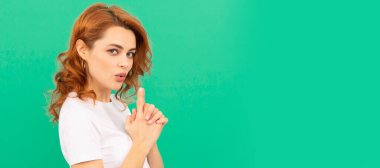 serious confident redhead girl with curly hair with gun gesture on blue background, confidence. Woman isolated face portrait, banner with copy space