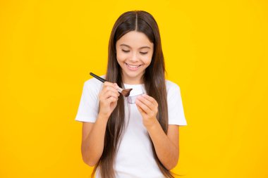 Güzellik, makyaj ve çocukların makyaj malzemeleri. Yüzüne allık süren genç bir kız sarı stüdyo arka planında izole edilmiş. Mutlu ergen portresi. Gülümseyen kız