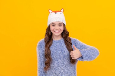 thumb up. positive kid with curly hair in hat. female fashion model. teen girl in knitwear on yellow background. portrait of child wearing warm clothes. express positive emotion. winter fashion.