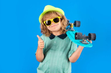 Studio portrait of stylish cute young child boy with skateboard isolated against blue background. Kid skater. Fashion kid. Stylish little child wearing a summer clothes, sunglasses and hat