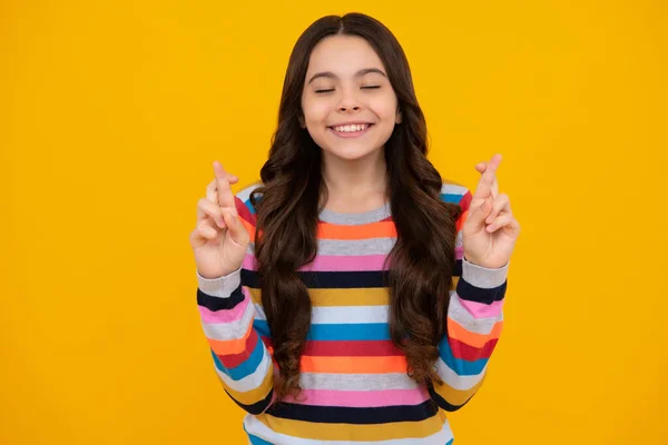 Teenager child holding fingers crossed for good luck. Portrait of cheerful girl prays and hopes dreams, crosses fingers for good luck, closes eyes, isolated on yellow studio background