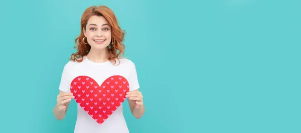 smiling redhead woman hold red heart on blue background. Woman isolated face portrait, banner with copy space