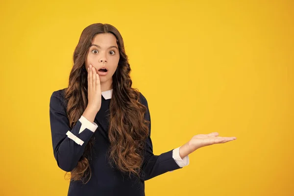 Surprised emotions of young teenager girl. Close up portrait of teenager child girl showing at copy space, pointing to ads advertising, isolated over yellow background