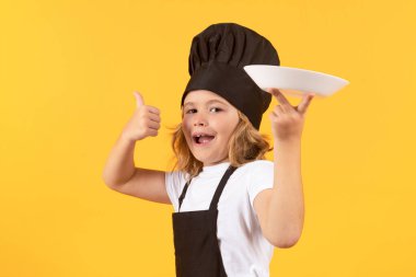 Funny child cook with cooking plate. Kid chef cook prepares food on isolated studio background. Kids cooking. Teen boy with apron and chef hat preparing a healthy meal