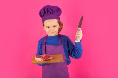 Child cook hold cutting board with meat beef steak and knife. Teen boy with apron and chef hat preparing a healthy meal