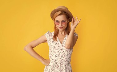 Sarı arka planda hasır şapkalı ve güneş gözlüklü gülümseyen kadın..