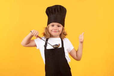 Funny kid chef cook with kitchen ladle, studio portrait. Kid chef cook prepares food on isolated studio background. Kids cooking. Teen boy with apron and chef hat preparing a healthy meal