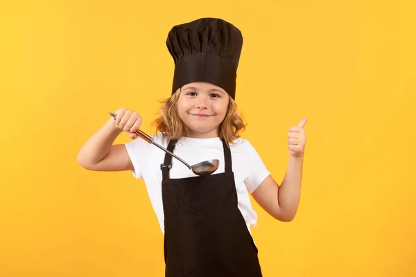 Funny kid chef cook with kitchen ladle, studio portrait. Kid chef cook prepares food on isolated studio background. Kids cooking. Teen boy with apron and chef hat preparing a healthy meal