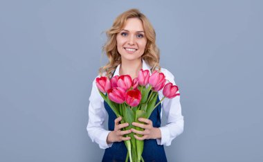 smiling young woman in apron with spring tulip flowers on grey background.