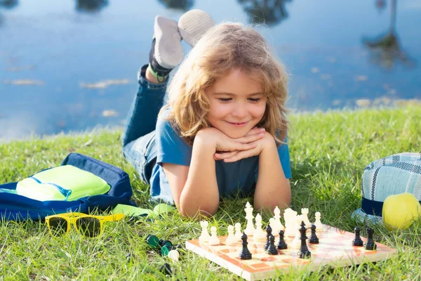 Concentrated child boy developing chess strategy, playing board game in backyard, laying on grass
