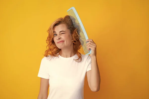 Surprised amazed girl combing hair. Redhead woman with a comb, isolated on yellow background. Woman brushing straight natural hair with comb. Girl combing hair hairbrush. Hair care beauty concept