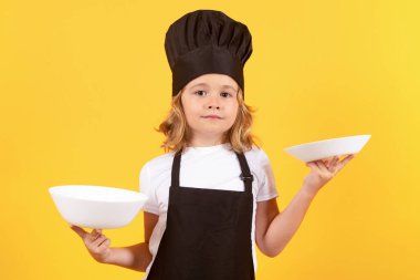 Little cook with cooking plate. Child in cook uniform. Chef kid isolated on yellow background. Cute child to be a chef. Child dressed as a chef hat