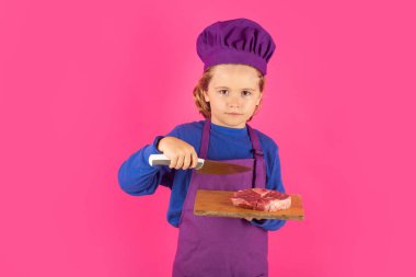 Child cook hold cutting board with meat beef steak and knife. Cooking, culinary and kids. Little boy in chefs hat and apron on studio isolated background