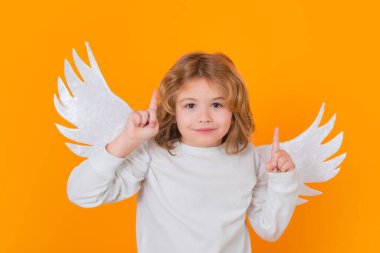 Child angel pointing up, point gesture, idea. Valentines day. Blonde cute child with angel wings on a yellow studio background. Happy angel child