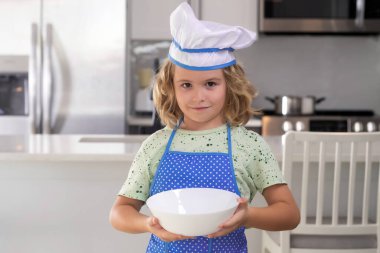 Child chef cook with cooking plate. Funny child stand at kitchen table have fun baking, doing bakery preparing food at home kithen