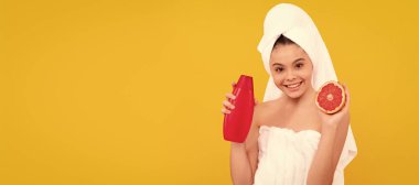 smiling kid in towel with grapefruit shampoo bottle on yellow background. Cosmetics and skin care for teenager child, poster design. Beauty kid girl banner with copy space