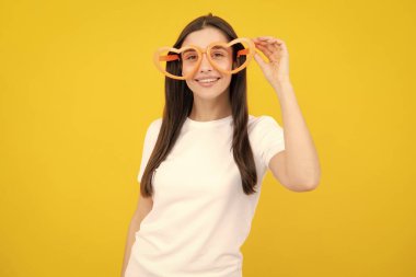 Kalp şeklinde gözlükleri olan, sarı stüdyo arka planında gülümseyen bir kız.