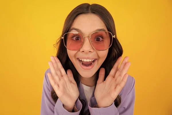 Amazed teen girl. Excited expression, cheerful and glad. Headshot portrait of cute teenager child girl isolated on yellow studio background wear sunglasses look at camera