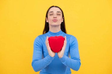 Mutlu kadın kırmızı kalbi tutar. İzole edilmiş sarı arka planda poz veren güzel bir kadın model. Neşeli, pozitif, tatlı, yumuşak kalpli bir kız.