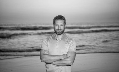 Sabah kumsalında traşsız gülümseyen adam. Gündoğumu kumsalında olgun bir adam. Gömlekli sakallı adam. Yaz modası. Yaz tatili. pozitif duygular. Deniz üzerinde gün batımında sakallı bir adam.