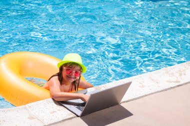 Çocuk yüzme havuzunda dizüstü bilgisayarla çalışıyor. Yaz çevrimiçi teknoloji. Yaz tatilinde tropikal suda dinlenen bir gezgin. Seyahat, dizüstü bilgisayar kullanan çocuk.