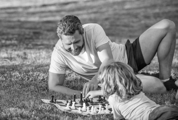 happy family of daddy and son boy playing chess on green grass in park outdoor, chessboard.