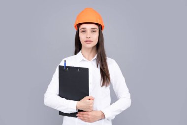 Serious woman worker in protective helmet with clipboard isolated on grey background. Young woman construction manager. Architect woman, female worker in hardhat helmet