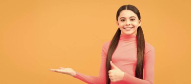 Portakal rengi arka planda fotokopi çeken mutlu genç kız başparmağını kaldır ve ürün teklifini yap. Çocuk yüzü, yatay poster, izole edilmiş genç kız portresi, fotokopi alanı olan pankart